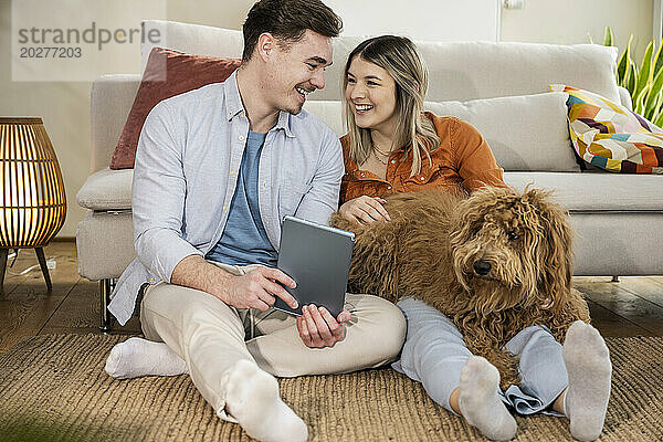 Glückliches junges Paar sitzt mit Tablet-PC und Hund zu Hause