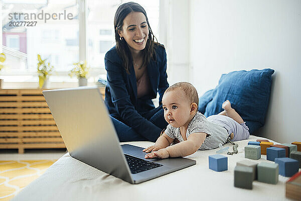 Glückliche Geschäftsfrau sitzt neben ihrem kleinen Mädchen und spielt mit Laptop im Bett