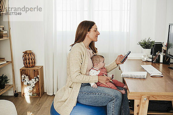 Berufstätige Frau sitzt mit Baby und hält Smartphone am Schreibtisch