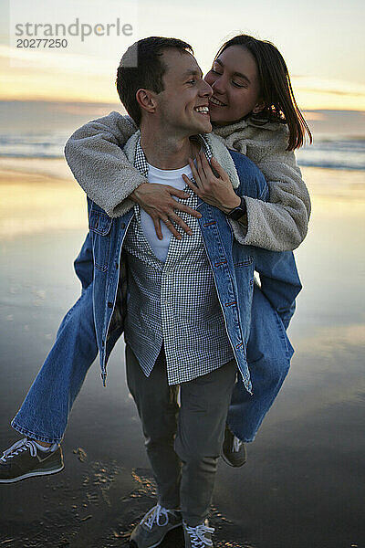 Happy boyfriend carrying woman piggyback on beach at sunset