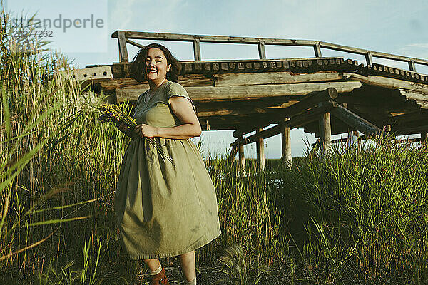 Lächelnde Frau  die zwischen Schilf in der Nähe einer Brücke im Feld spaziert