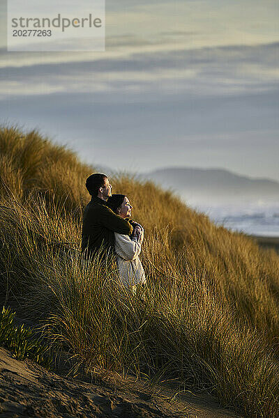 Mann umarmt Freundin von hinten und steht in der Düne am Strand
