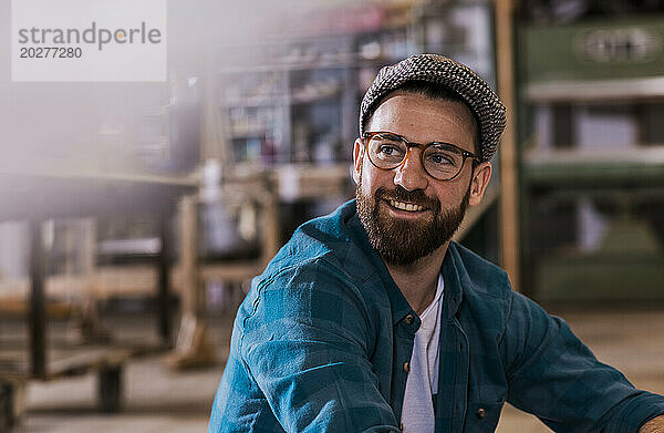 Lächelnder Tischler mit Brille und Schiebermütze in der Werkstatt