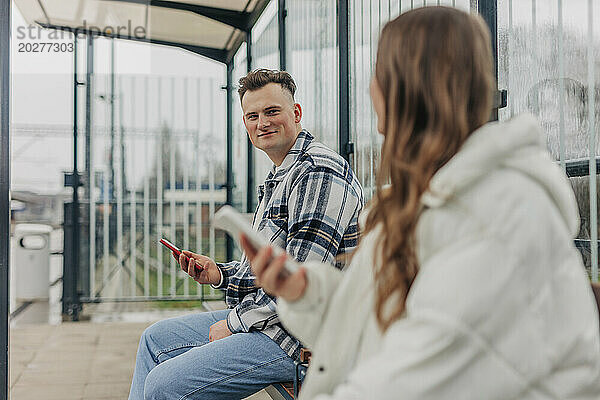 Junges Paar sitzt auf einer Bank und hält Smartphones am Bahnhof