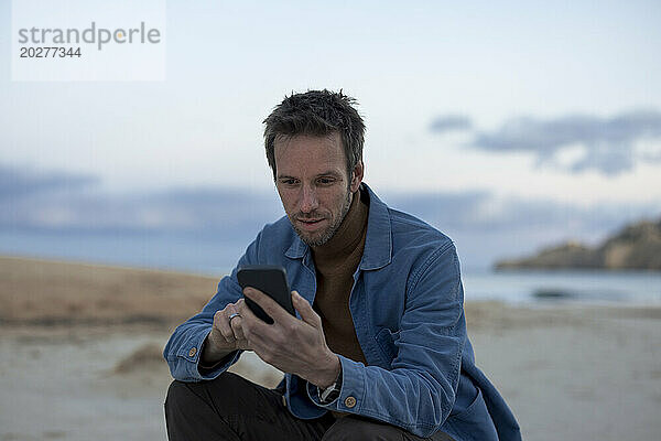 Mann benutzt Smartphone am Strand bei Sonnenuntergang