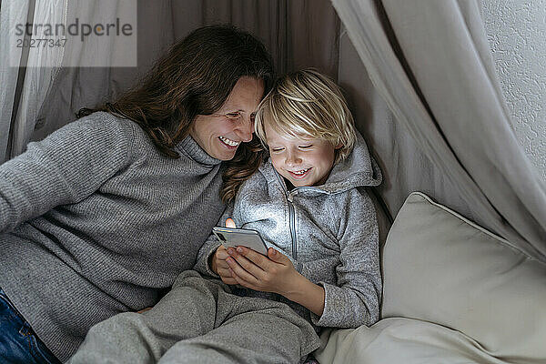 Glückliche Mutter blickt ihren Sohn mit seinem Smartphone im grauen Baldachin an