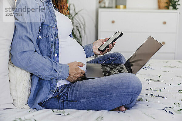 Schwangere Frau sitzt mit Smartphone und Laptop im Heimbüro im Bett