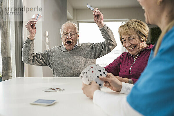 Fröhlicher Mann spielt Karten mit Frau und häuslicher Pflegekraft am Tisch