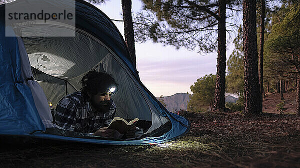 Mann mit Stirnlampe und Lesebuch im Zelt bei Sonnenuntergang