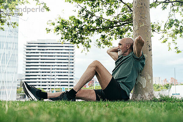 Retired senior man leaning on tree trunk with hands behind back at park