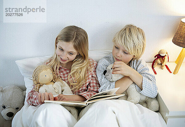 Smiling girl reading book with brother sitting on bed at home