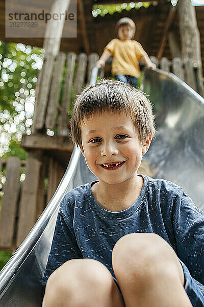 Happy boy sliding down on slide with brother in background at playground