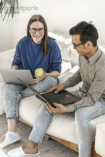 Lächelnde  vielfältige Programmierer mit Laptops sitzen auf dem Sofa im kreativen Startup-Büro