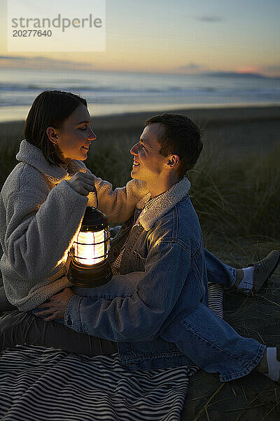 Lächelndes Paar mit Laterne verbringt gemütlichen Sonnenuntergang am Strand