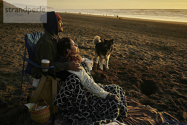 Junges Paar entspannt sich bei Sonnenuntergang am Strand mit Hund