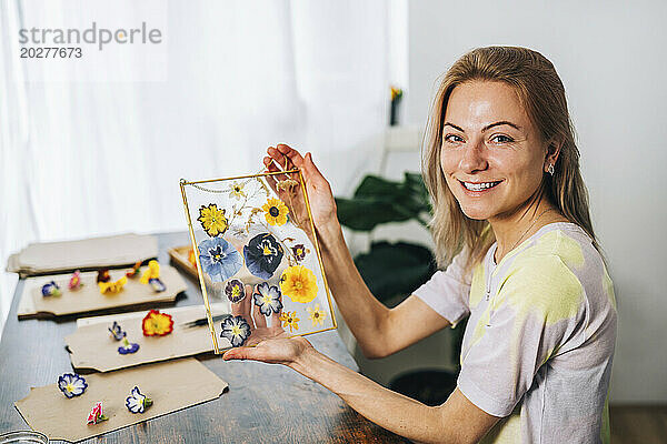 Glückliche Frau hält zu Hause einen transparenten Rahmen  der mit Trockenblumen geschmückt ist