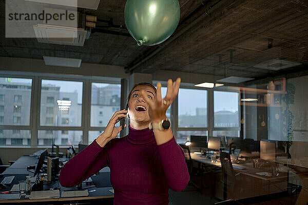 Playful businesswoman talking on smart phone and catching balloon in office