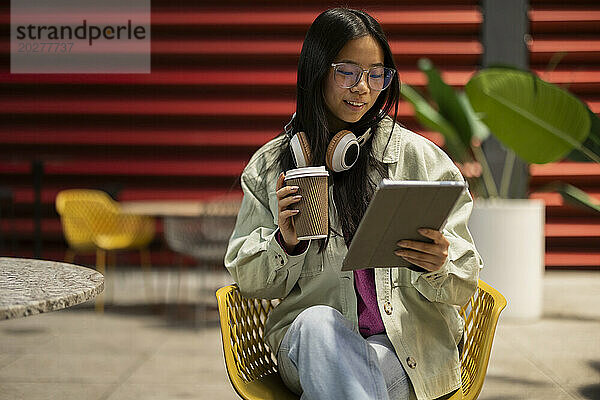 Lächelnde junge Frau hält Kaffeetasse in der Hand und benutzt Tablet-PC im Straßencafé