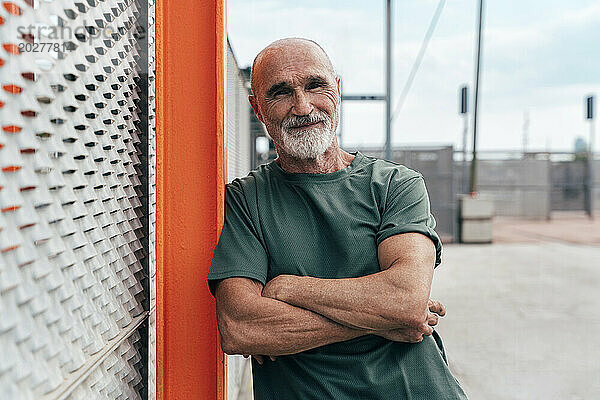 Lächelnder  aktiver älterer Mann mit verschränkten Armen  der sich an den Zaun lehnt