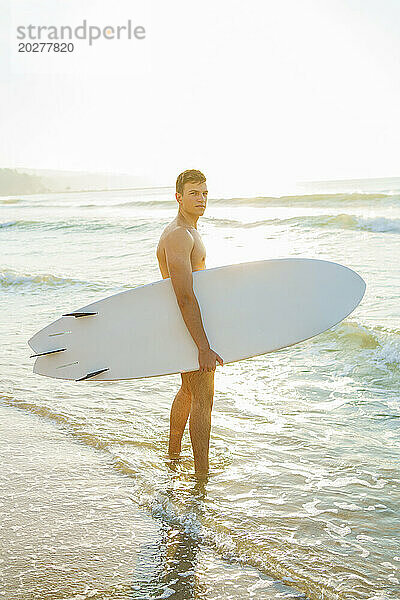 Junger Mann mit Surfbrett steht bei Sonnenuntergang im Wasser