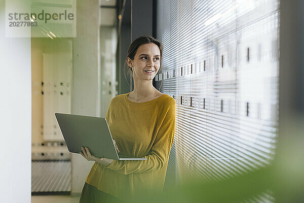 Glückliche junge Geschäftsfrau steht mit Laptop im Büroflur