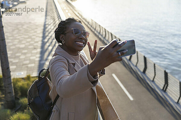 Lächelnde Frau gestikuliert ein Friedenszeichen und macht ein Selfie am Fluss