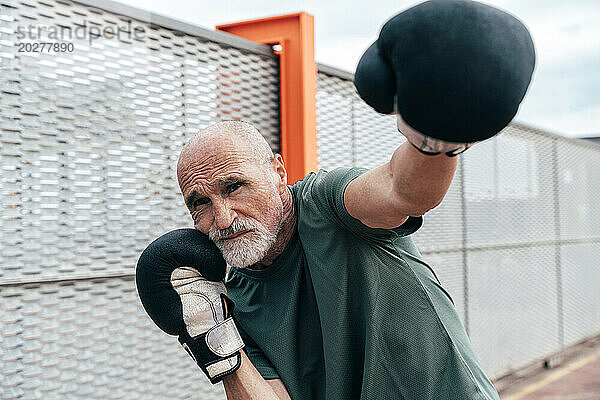 Selbstbewusster älterer Mann übt mit Boxhandschuhen am Zaun