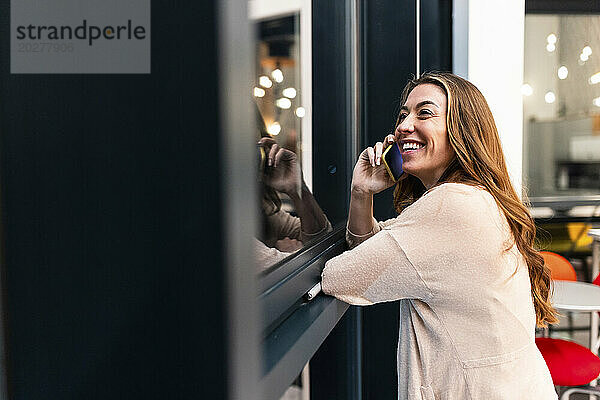 Glückliche Geschäftsfrau  die am Fenster im Büro mit dem Smartphone spricht