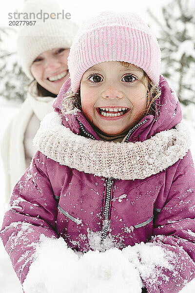 Aufgeregtes Mädchen hält Schnee mit Mutter im Hintergrund im Winterpark