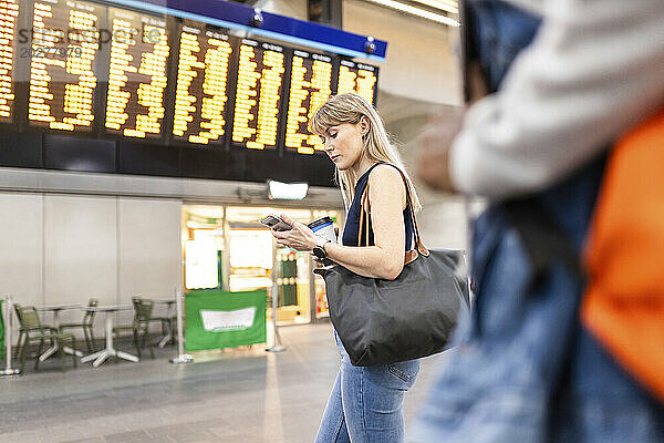 Frau benutzt Smartphone und steht in der Nähe der Ankunfts- und Abfahrtstafel am Bahnhof