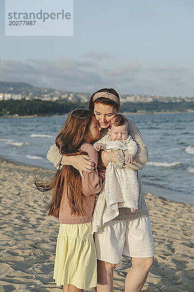 Glückliche Mutter umarmt ihre Töchter an einem sonnigen Tag am Strand