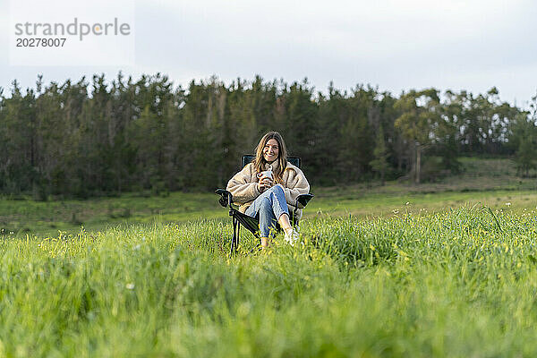 Lächelnde Frau sitzt auf einem Campingstuhl und hält eine Kaffeetasse auf der Wiese
