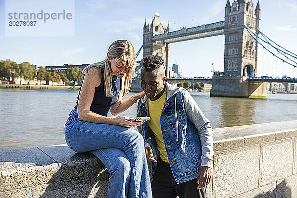 Lächelnde Frau teilt Smartphone mit Freund in der Nähe der Tower Bridge in der Stadt