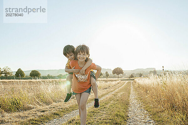 Smiling girl piggybacking brother in field at sunset