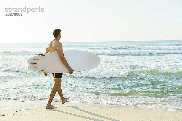 Mann läuft mit Surfbrett am Strand in Richtung Meer