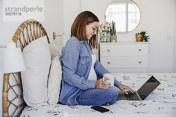Schwangere Freiberuflerin hält Tasse und Laptop im Bett im Heimbüro