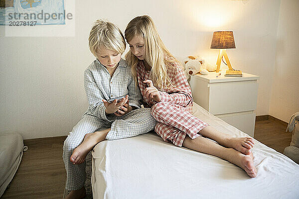 Geschwister im Schlafanzug sitzen mit Smartphone zu Hause im Bett