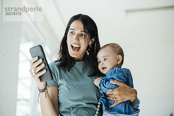 Fröhliche Mutter benutzt Telefon und trägt ihre kleine Tochter im heimischen Wohnzimmer