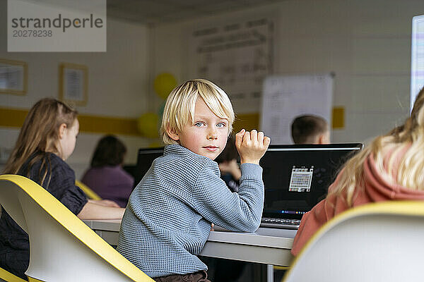 Blonder Junge blickt über die Schulter zurück und sitzt mit Laptop am Schreibtisch im Klassenzimmer