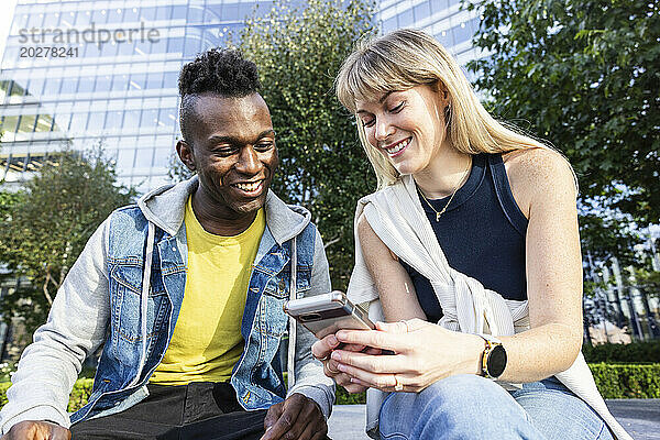 Lächelnde  gemischtrassige Freunde teilen sich ihr Smartphone und sitzen in der Nähe des Gebäudes