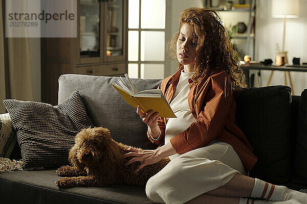 Expectant woman reading book sitting with dog on sofa in living room