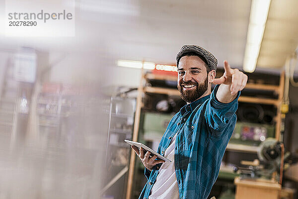 Happy carpenter holding tablet PC and pointing at workshop