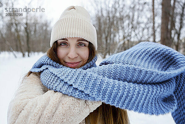 Lächelnde Frau mit Strickmütze und blauem Schal im Winterwald