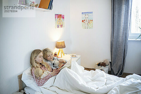 Blond brother and sister reading book on bed at home