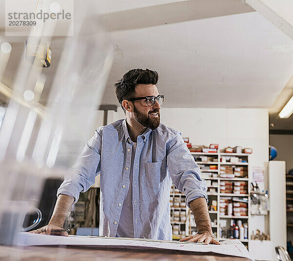 Lächelnder Handwerker lehnt in der Werkstatt am Tisch