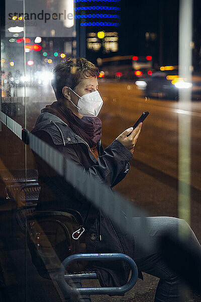 Reife Frau trägt eine schützende Gesichtsmaske und benutzt ihr Mobiltelefon  während sie an einer Bushaltestelle in der Stadt sitzt
