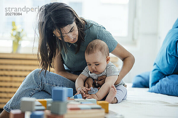 Mother sitting by baby daughter playing with toys on bed at home