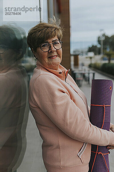 Ältere Frau mit Gymnastikmatte lehnt an Glaswand