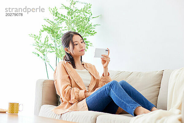 Asiatische Frau sitzt auf der Couch und hält eine Kaffeetasse