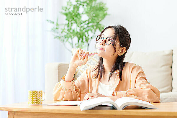 Asiatische Frau mit Brille sitzt mit Buch und Stift am Tisch